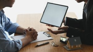 Cropped shot of property agent seller in formal suit explaining about policy/agreement to his customer by using tablet at the wooden desk. House model and clipboard on the wooden desk as background. After having a home inspection.