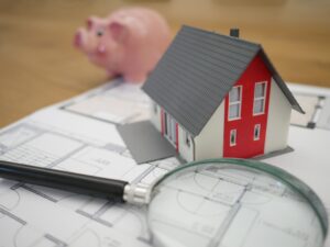 Picture of objects spread over a table. A magnify glass, 3D small house, house plans, and a piggy bank.